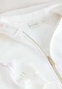 Vêtement blanc zippé avec une fermeture éclair dorée, marquage Baker sur l'étiquette du col et la manche, et un petit motif clé rose sur la manche.