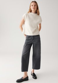 Vrouw in een getextureerde witte mouwloze top, met korte wijde zwarte jeans en zwarte instappers, staand met handen in haar achterzakken.