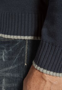 Marineblauer Strickpullover mit geripptem Saum, der hellgraue Akzente aufweist; kombiniert mit abgetragenen dunkelblauen Jeans, die kontrastierende Steppnähte zeigen.