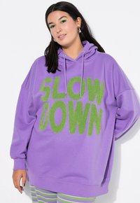 Fioletowy, oversize'owy hoodie z zielonym, puszystym napisem "SLOW DOWN"; posiada kaptur ze sznurkiem i mankiety z długimi rękawami. Widoczne paskowane spodnie poniżej.