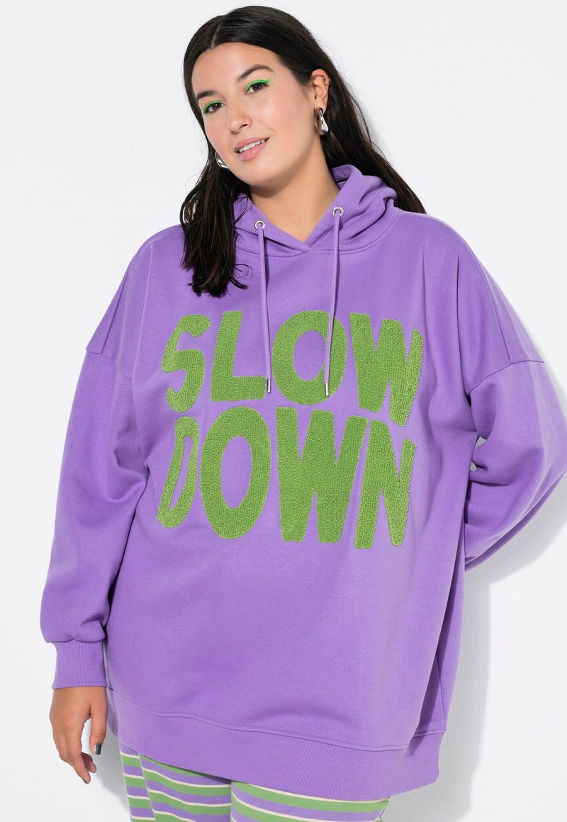 Fioletowy, oversize'owy hoodie z zielonym, puszystym napisem "SLOW DOWN"; posiada kaptur ze sznurkiem i mankiety z długimi rękawami. Widoczne paskowane spodnie poniżej.