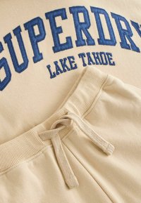 Béžová mikina s modrým vyšitým nápisom "SUPERDRY LAKE TAHOE" a béžové tepláky so sťahovacou šnúrkou, ribbed pásom a hladkou textúrou.