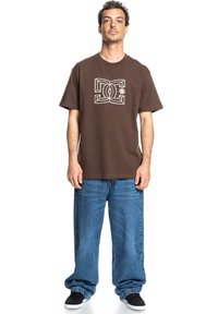 Camiseta de algodón marrón con un diseño gráfico blanco en el frente. Llevada con unos pantalones vaqueros de algodón azul sueltos y zapatos negros.