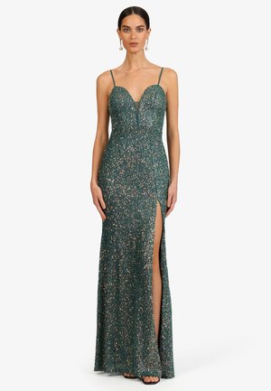 Frau trägt ein bodenlanges dunkelgrünes glitzerndes Kleid mit dünnen Trägern, tiefem Ausschnitt und hohem Beinschlitz, stehend vor einem schlichten Hintergrund.