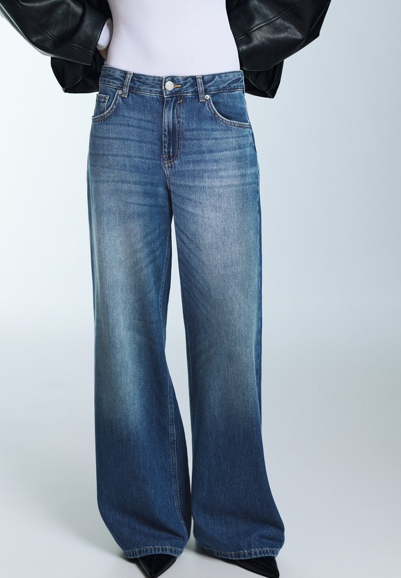 Modré džíny s širokými nohavicemi vyrobené z denimu, s vysokým pasem, předními kapsami a vybledlým vzhledem, doplněné černými špičatými botami.