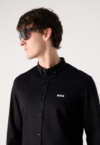 Zwarte langetermijnhemd met knoopsluiting en button-down kraag, met een witte "BOSS" logo aan de linkerkant en glanzende zwarte knopen. Model draagt een zonnebril.