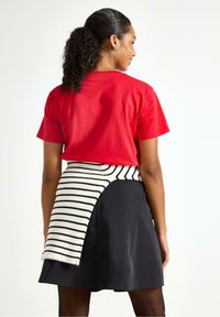 Czerwona koszulka z krótkim rękawem w połączeniu z czarną spódnicą, która ma białą i czarną paskowaną tkaninę owiniętą wokół, tworząc efekt warstwowy.