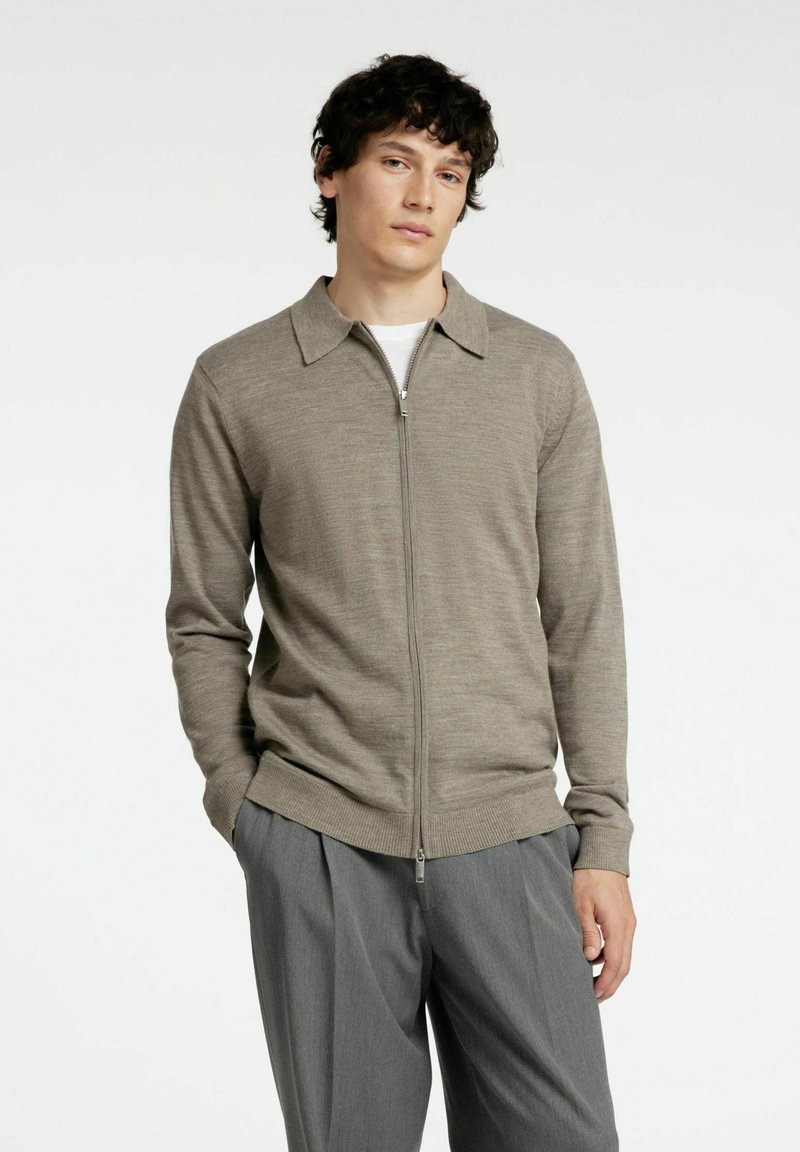 Svetlo rjava zip-up pulover s ovratnikom, rebrastimi manšetami in robom. Uporabljen preko bele majice, v kombinaciji s sivo krilom. Gladka tekstura.