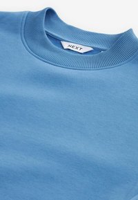Világoskék pulcsi, bordázott kerek nyakkal, sima textúrával, és a „NEXT” márkát jelző címkével a nyakán.