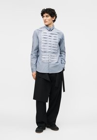 Homme portant une chemise bleue à boutons avec des détails en tissu plissé horizontal et un pantalon noir à jambes larges avec un voile asymétrique.