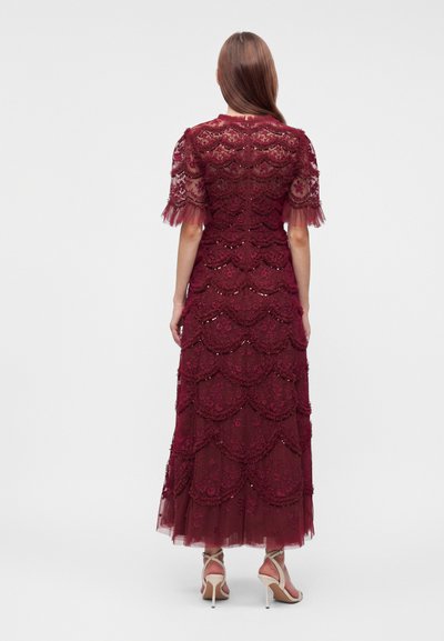 Femme portant une longue robe en dentelle bordeaux avec des couches festonnées, des manches courtes et des détails transparents, associée à des sandales blanches à talons hauts.
