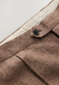 Pantalones marrones texturizados con un forro interior suave y un detalle de botón negro. Presentan pliegues marcados y elementos de diseño a medida.