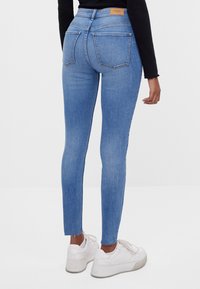 Jeans ajustados de denim azul medio; cuentan con una cintura alta, dos bolsillos en la parte posterior y un dobladillo deshilachado. Combinados con zapatillas blancas.