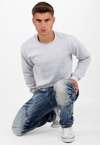 Sweatshirt en polaire gris, jeans bleus déchirés avec des accents au niveau des genoux, et baskets blanches avec des bords rouges. Le mannequin est à genoux avec les mains sur le genou.
