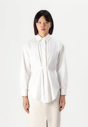Camisa blanca de botones con un cuerpo entallado, dobladillo curvado y mangas largas. Presenta un detalle de torsión en el centro y una textura de tela suave.