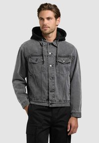 Jeune homme portant une veste en jean noire délavée par-dessus un sweat à capuche noir, assorti à un pantalon cargo noir, debout devant un fond uni.