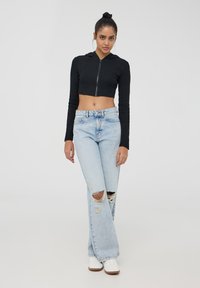 Cappuccio nero corto con zip abbinato a jeans sbiaditi a gamba larga blu chiara con strappi alle ginocchia, abbinato a sneakers bianche.