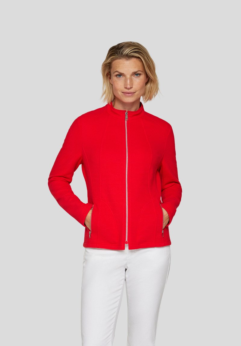 Red zip-up jacket with a textured pattern, high collar, and side pockets, paired with white pants.
