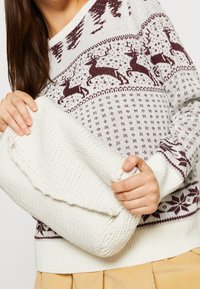 Pull en tricot crème avec motifs de vacances bordeaux, comprenant des rennes et des motifs géométriques. Le modèle tient une pochette blanche texturée.