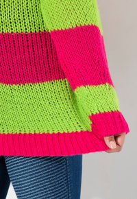 Pull en tricot avec des rayures horizontales en rose vif et vert. Coupe ample, poignets côtelés et fil texturé avec un tissage ouvert bien visible.