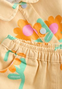 Tissu jaune avec ceinture élastique présentant des fleurs orange et roses avec des tiges vertes et des boutons-pression argentés sur un vêtement.