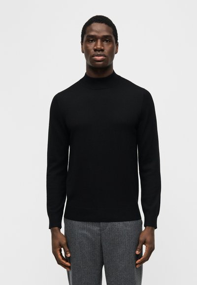 Άνδρας που φοράει μαύρο πουλοβεράκι με ψηλό γιακά και γκρι παντελόνι, στέκεται μπροστά σε απλό φωτεινό φόντο.