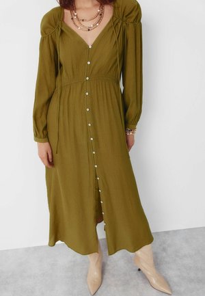 Kvinde iført en olivengrøn midi-kjole med knapper foran og lange ærmer, lag-på-lag halskæder, armbånd og beige ankelstøvler med spids tå.