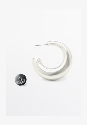 Zilveren metalen hoepeloorbel met een glad, gebogen ontwerp. Heeft een slanke steker en een ronde, beveiligde achterkant met merkdetails.