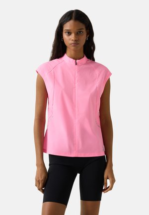 Kvinde iført en ærmeløs lyserød top med lynlås og sorte cykelshorts, stående mod en ensfarvet lys baggrund.