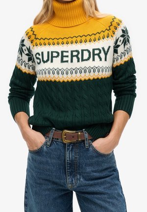 Mujer lleva un suéter de punto con cuello alto en verde, blanco y amarillo con texto "SUPERDRY" y diseño estampado, combinado con jeans azules y cinturón marrón.