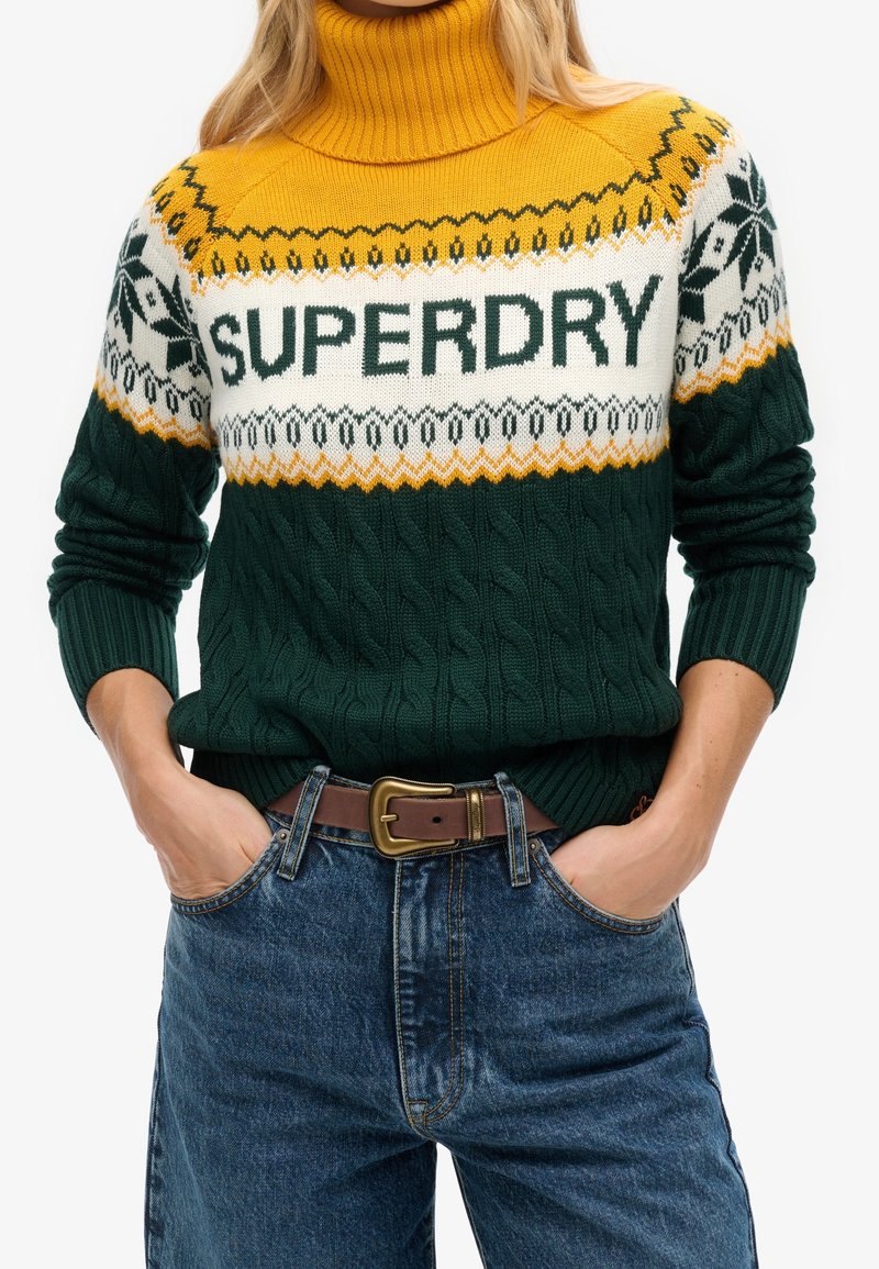 Une femme porte un pull à col roulé tricoté vert, blanc et jaune avec le texte "SUPERDRY" et un motif, assorti à un jean bleu et une ceinture marron.