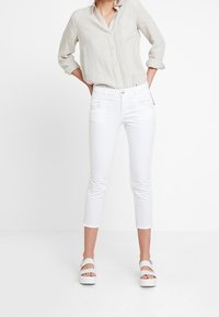 Mujer vistiendo una camisa larga de manga beige, jeans ajustados blancos cortos y sandalias de plataforma blancas, de pie con las manos en las caderas.