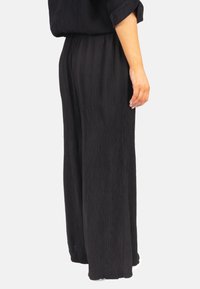 Pantalons larges plissés noirs en tissu texturé, avec une taille élastique haute et une silhouette fluide. Met en valeur la facilité de mouvement.