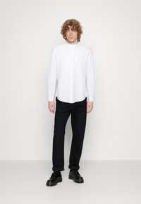 Witte langbloes met knoopsluiting en een bandkraag, gecombineerd met zwarte broek met opgerolde manchetten en zwarte veterschoenen.
