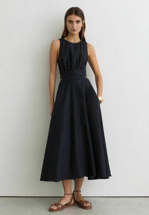 Młoda kobieta w czarnej, bezrękawowej sukience midi z wycięciami w talii, brązowych sandałach i złotej bransoletce, stojąca na tle białej ściany.