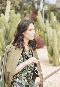 Green patterned shirt with leafy designs, olive jacket, gold necklace, holding white sunglasses. Desert background with cacti.
