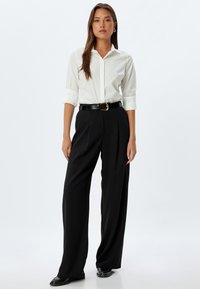 White button-up shirt with rolled sleeves, black wide-leg trousers, cinched with a glossy black belt. Model standing against a plain background.