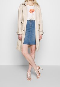 Beige trenchcoat over en hvid blomster t-shirt, parret med en denimnederdel og hvide hælede sandaler. Looket kombinerer afslappede og klassiske elementer.