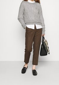 Pull gris tricoté, chemise blanche avec col, pantalon marron et mocassins noirs. Sac à main noir avec foulard à motifs drapé dessus.