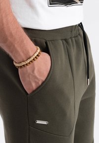 Olijfgroene joggingbroek met een zachte textuur, voorzien van een zijzak, elastische tailleband en een klein zilveren logo accent aan de voorkant.