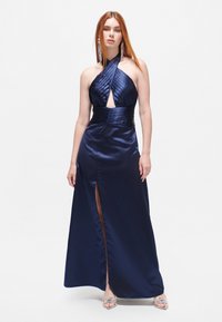 Satin halter gown navy dengan bodice berplis, celah samping, dan pinggang pas, dipadukan dengan sandal bertumit transparan.
