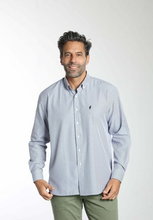 Homme aux cheveux foncés et barbe, portant une chemise rayée à boutons et un pantalon vert, debout devant un fond blanc uni.