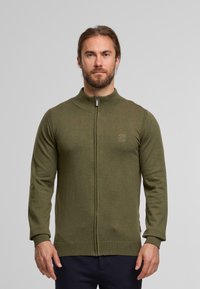 Cardigan zippé kaki en tissu tricot doux. Il dispose d'un col haut et de poignets côtelés, avec un petit logo sur la poitrine.