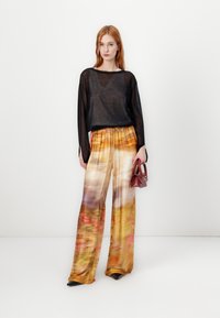 Svart genomskinlig långärmad topp med ljusgrå detaljer; vida byxor med ett flerfärgat, suddigt abstrakt mönster; håller en liten röd handväska.