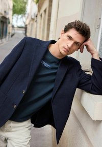 Blazer navy à revers crantés et deux boutons sur un t-shirt vert foncé avec du texte. Pantalon beige clair. Fond urbain doux.