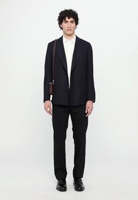 Blazer bleumarin cu două rânduri de nasturi, cămașă albă, pantaloni negri, pantofi negri și o geantă de umăr maro, purtate de un model în picioare pe un fundal alb.