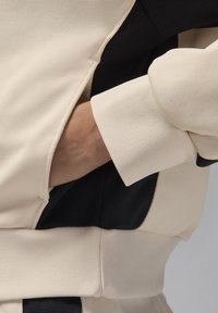 Tissu texturé beige et noir, avec un détail de poche et des manches à revers. Le design fusionne des textures lisses et côtelées.
