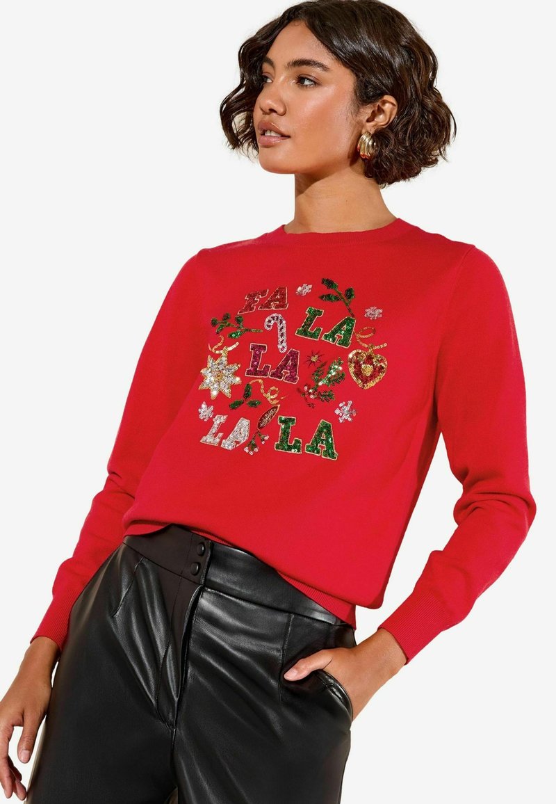 Rode trui met de tekst "FA LA LA" en kerstmotieven in het groen en goud. Lange mouwen, ronde hals, gedragen met zwarte leren broek.