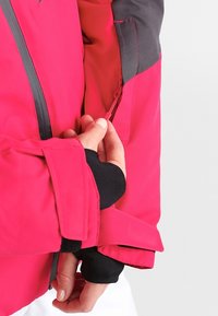 Giacca rosa brillante con un accento nero. Presenta una tasca con zip all'interno della manica sinistra e polsini elasticizzati per una vestibilità aderente.