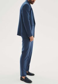 Mėlynas siuvinėtas kostiumas su tekstūruotu paviršiumi, sudarytas iš vienos eilės švarko ir siaurai prigludusių kelnių, derinamas su tamsiais loafers batais.